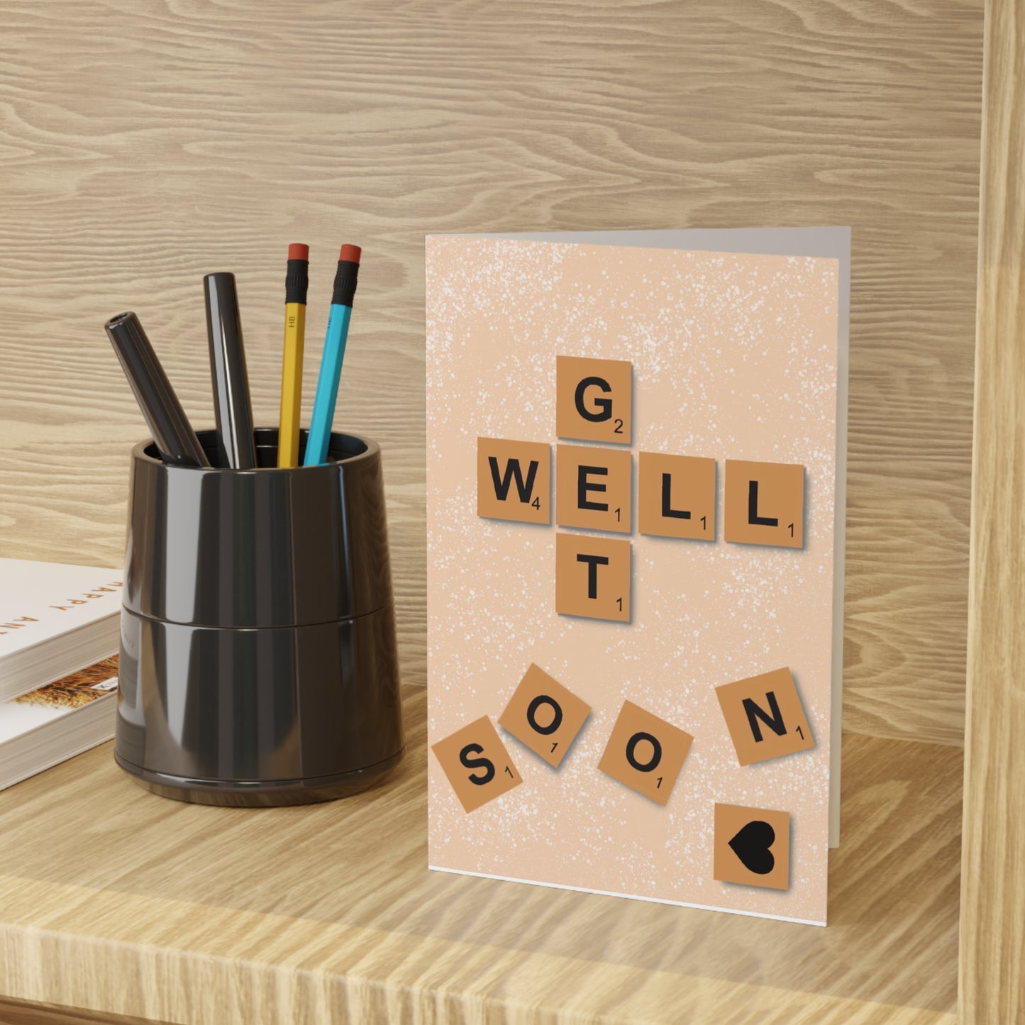 A beige speckled greeting card on a wooden surface has a pixelated finish with Scrabble tiles reading "GET WELL SOON" with a black heart tile at the bottom right. A black cup of pens is to the left.