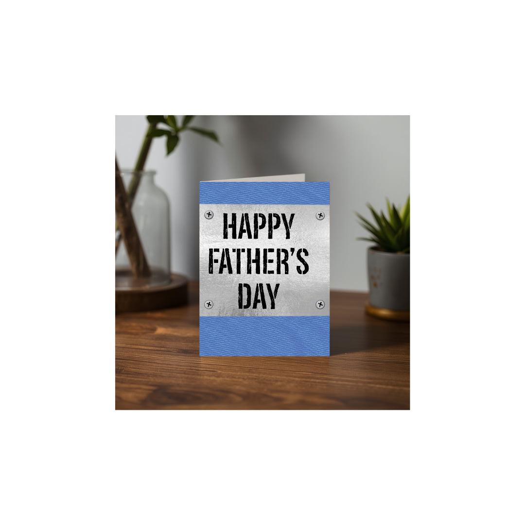 Blue denim Father's Day greeting card with metal sign design displayed on a wooden desk surrounded by green plants.