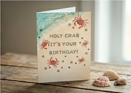 Holy crab it's your birthday' beach-themed card with crabs and starfish on sand styled on a wooden table with seashells