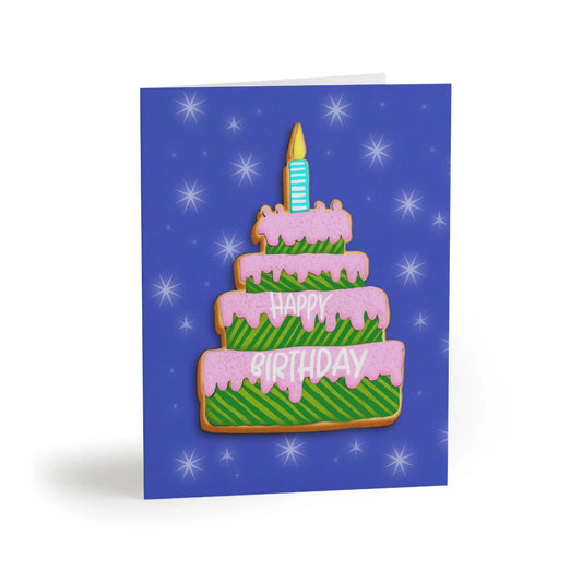 “A birthday card with a tiered cookie cake design, pink frosting, green striped cake, and cookie trim, on a blue star-filled background. The words "HAPPY BIRTHDAY" are on the cake.”