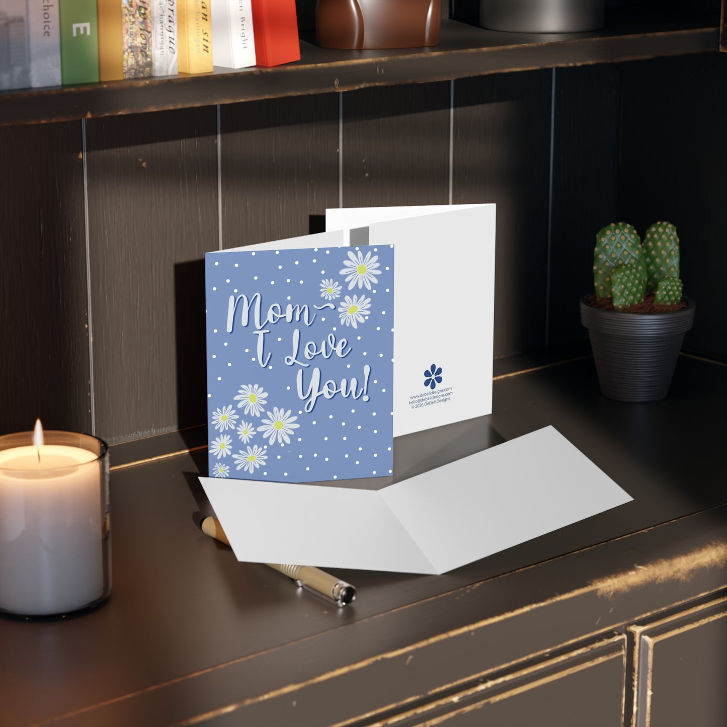 “A blue Mother's Day card with white polka dots and daisies, text "Mom I Love You!" and a white open card next to it, on a wooden desktop.”