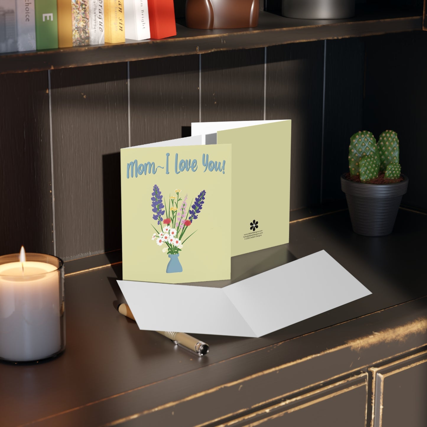 {"altText": "A light chartreuse greeting card with "Mom~ I Love You!" and an illustration of wildflowers in a blue vase, sitting open on a dark wooden surface with a lit candle, silver pen, and potted cactus."}