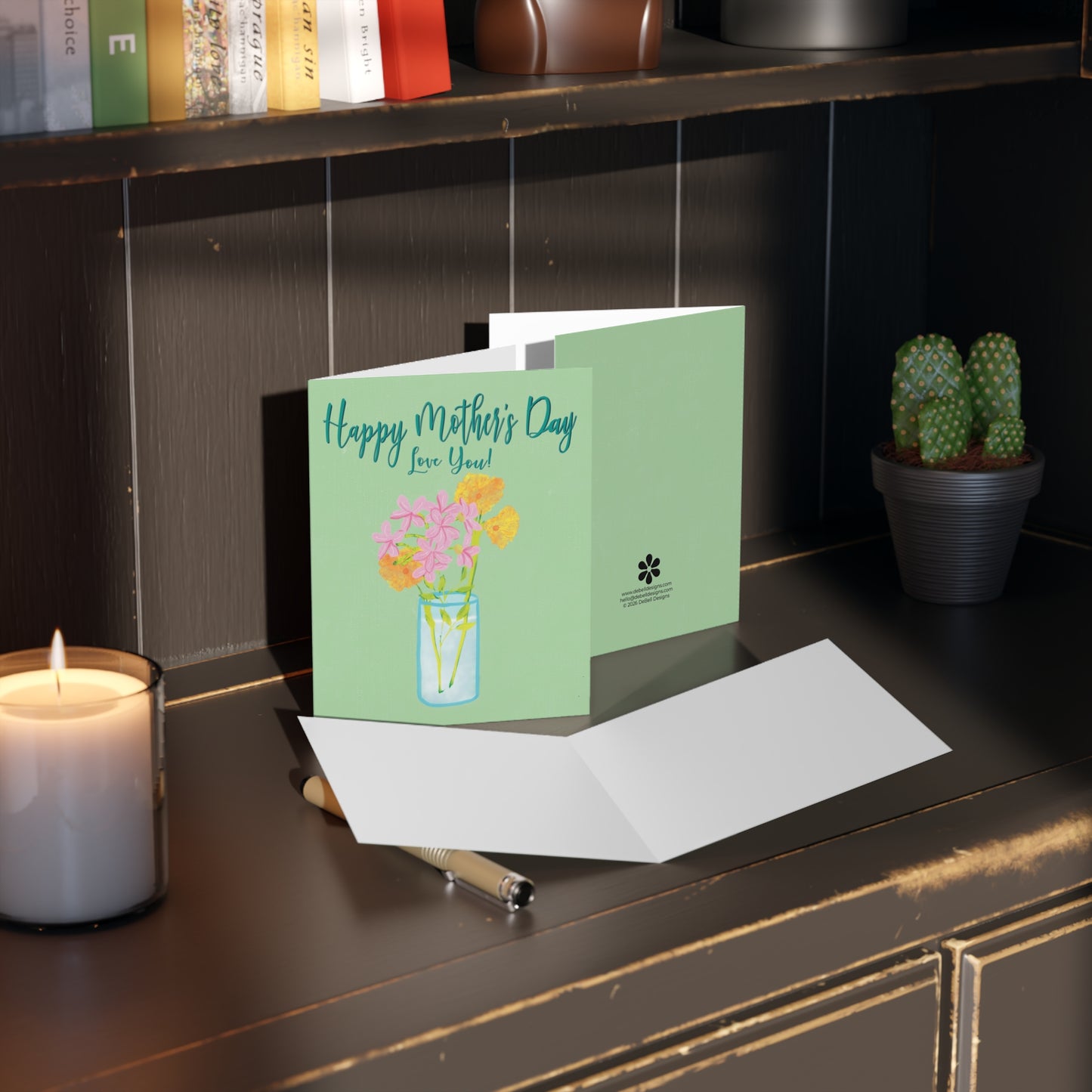 {"altText": "A light green greeting card with pink and yellow flowers in a vase reads, "Happy Mother's Day Love You!" on a dark brown desk next to a lit candle."}