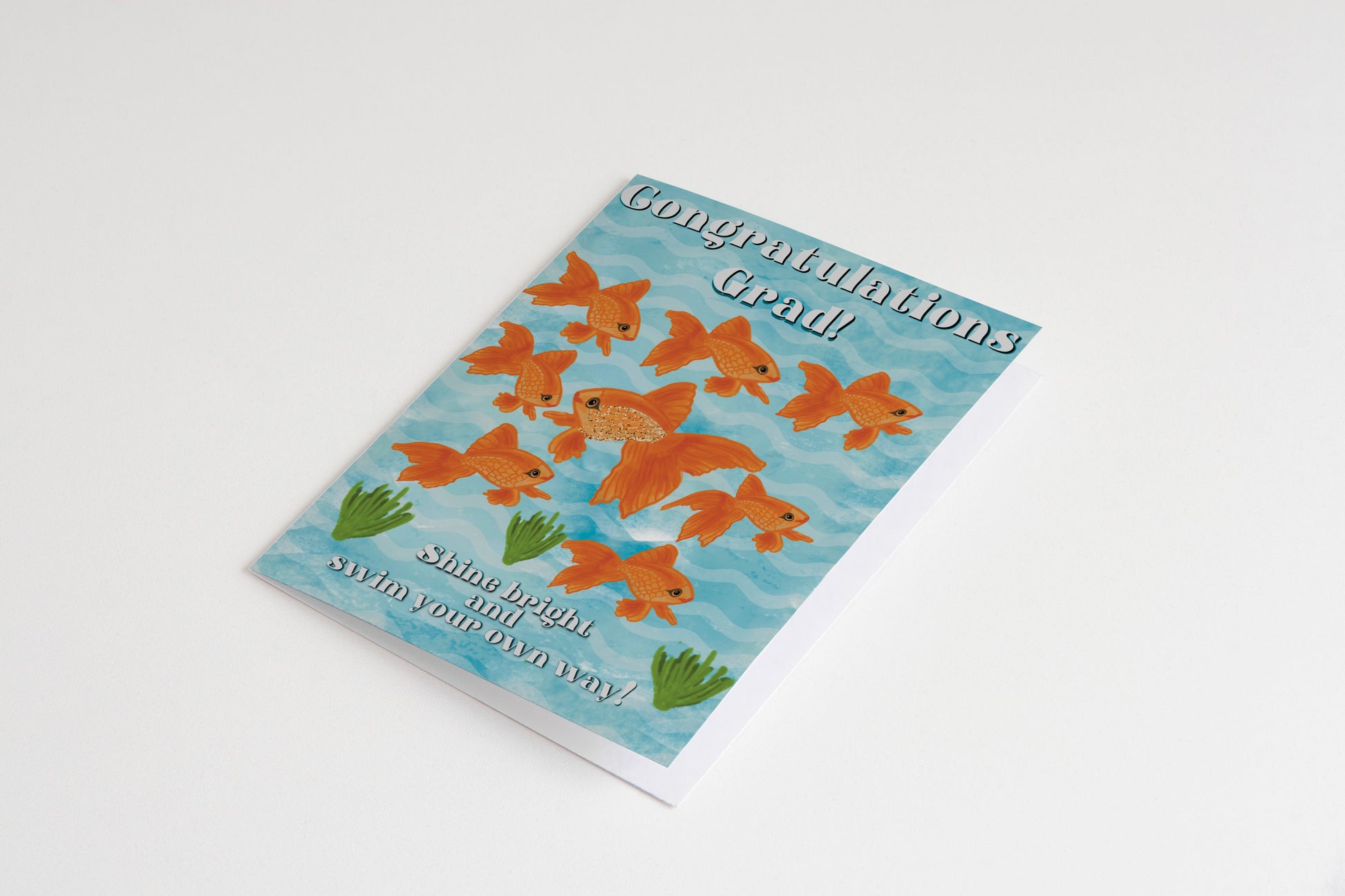 “Graduation card featuring 9 orange goldfish and aquatic plants against a light blue, wavy background. Text reads, "Congratulations Grad! Shine bright and swim your own way!”"