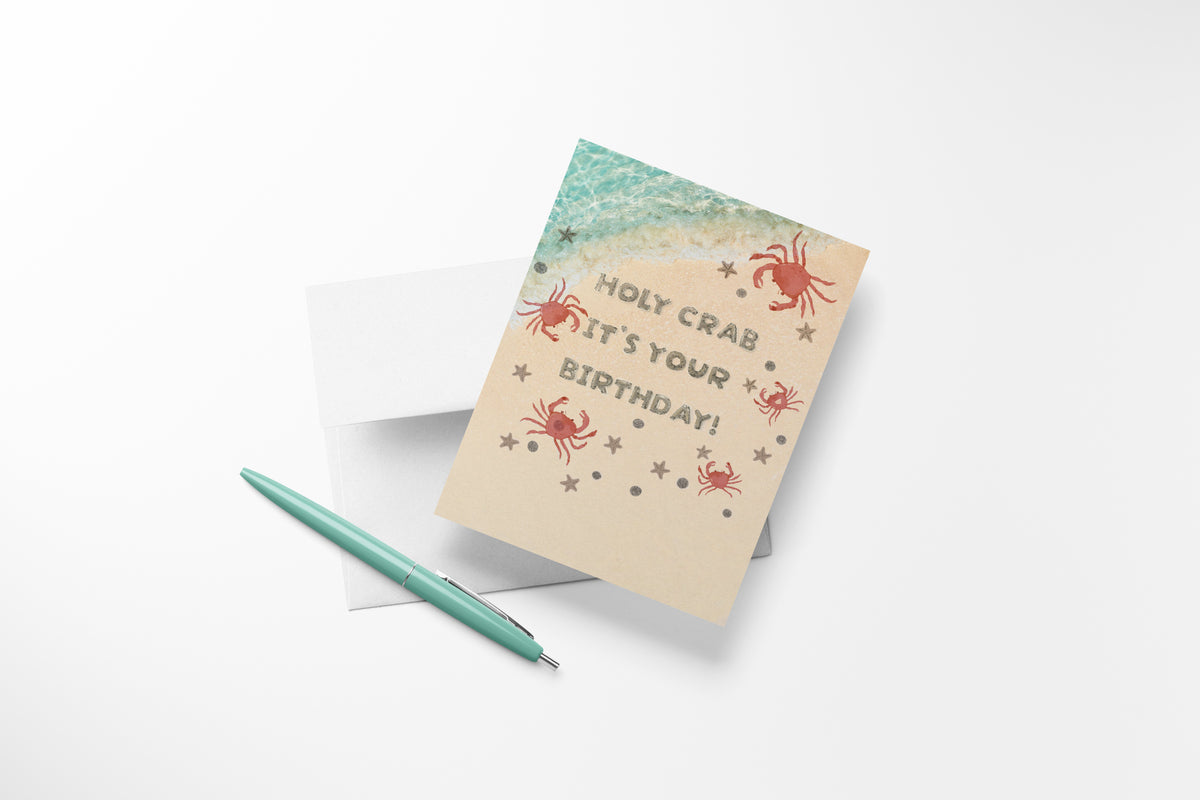 Holy crab it's your birthday' beach-themed card with crabs and starfish on sand with white envelope and sea green pen