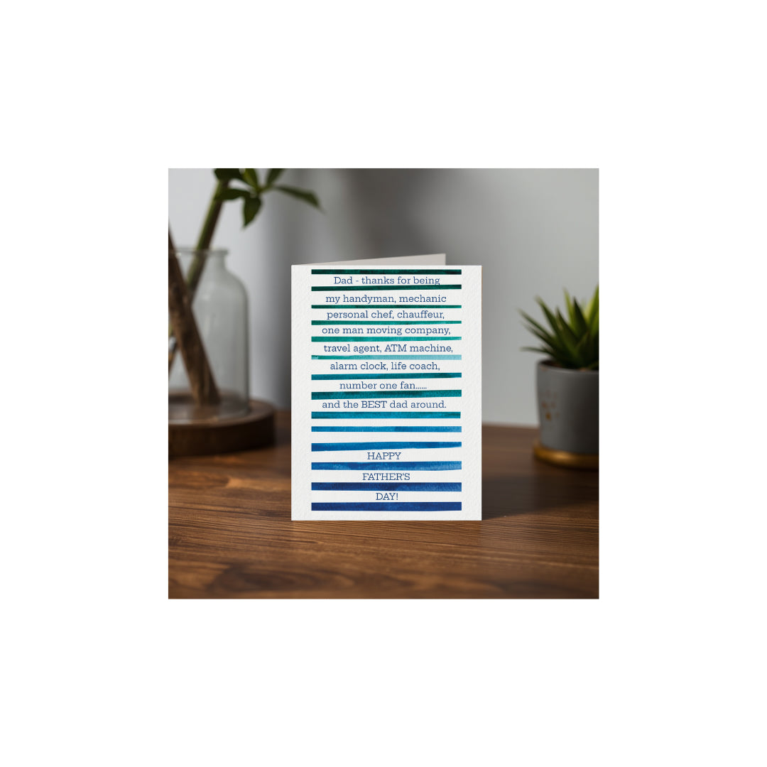Teal and navy watercolor stripe Father's Day card with a humorous sentiment displayed on a wooden desk with green plants.
