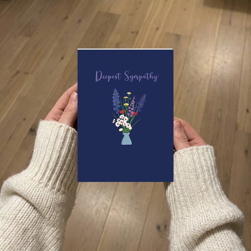A person holding a navy blue greeting card with the words 'Deeplest Sympathy' and a colorful hand-illustrated floral in the center, photographed from below over a light wood floor.
