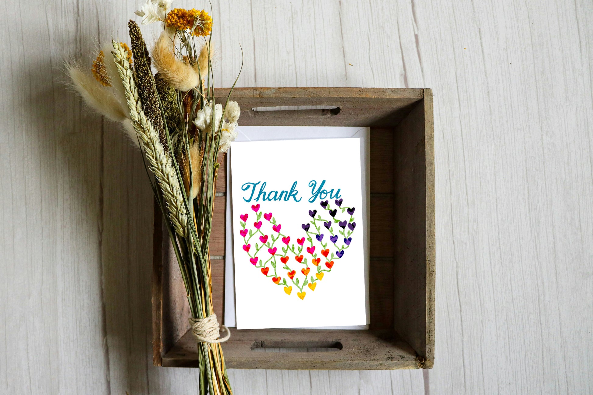 DeBell Designs rainbow hearts thank you greeting card displayed in a wooden tray with dried flowers, cozy lifestyle styling.