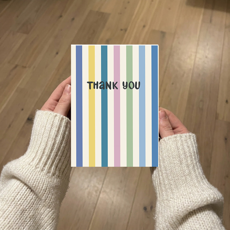 A person holding a vertical striped thank you card with pastel vertical stripes and the words 'THANK YOU' in black, photographed over a light wood floor background.